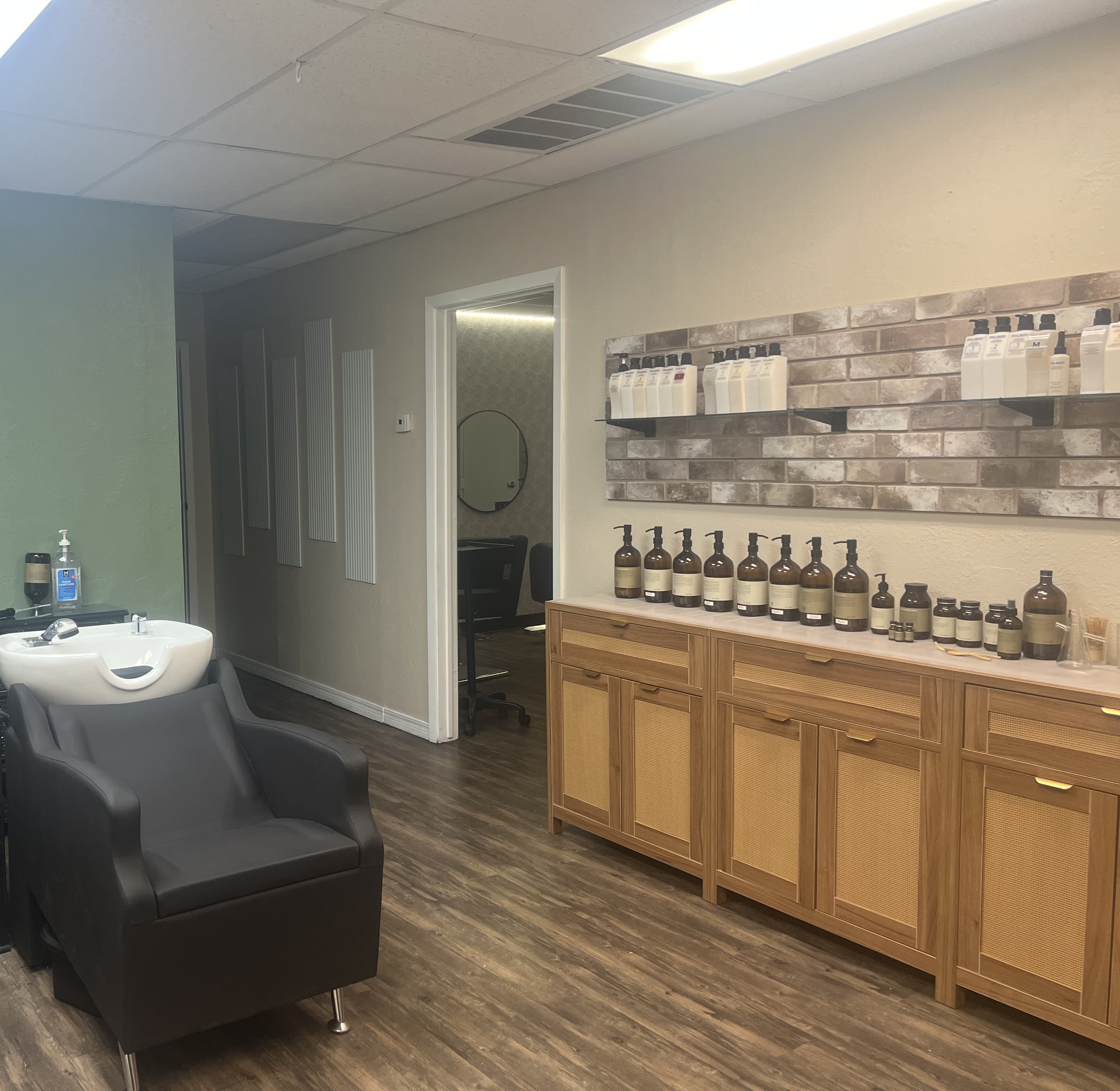 Shampoo bowl and a long cabinet with brickette and glass shelves on the wall with our Milbon Pro treatments and Oway head spa hair baths, powders, and oils